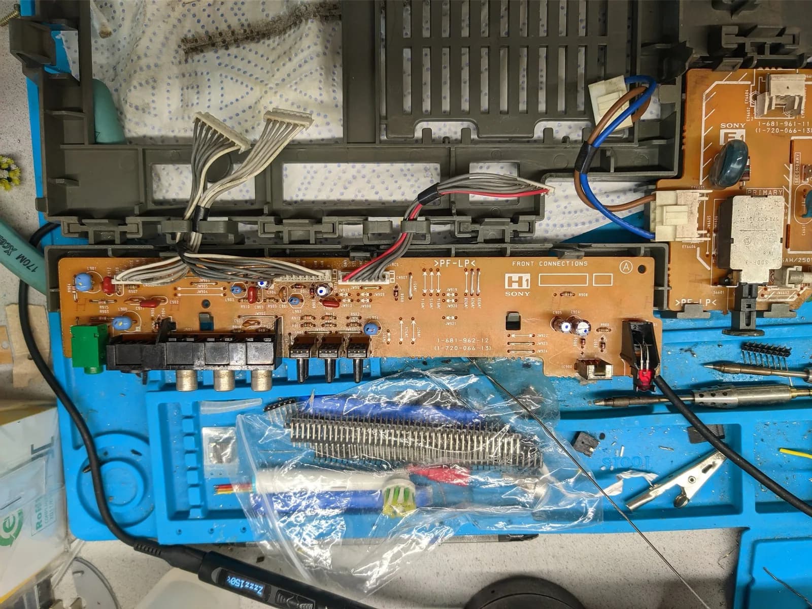 Sony KV-29FX30E front connections board (silkscreened FRONT CONNECTIONS, with RCA composite jacks) on a blue silicone mat, ribbon and power leads attached, a bagged strip of new parts and a soldering iron beside it during the recap.