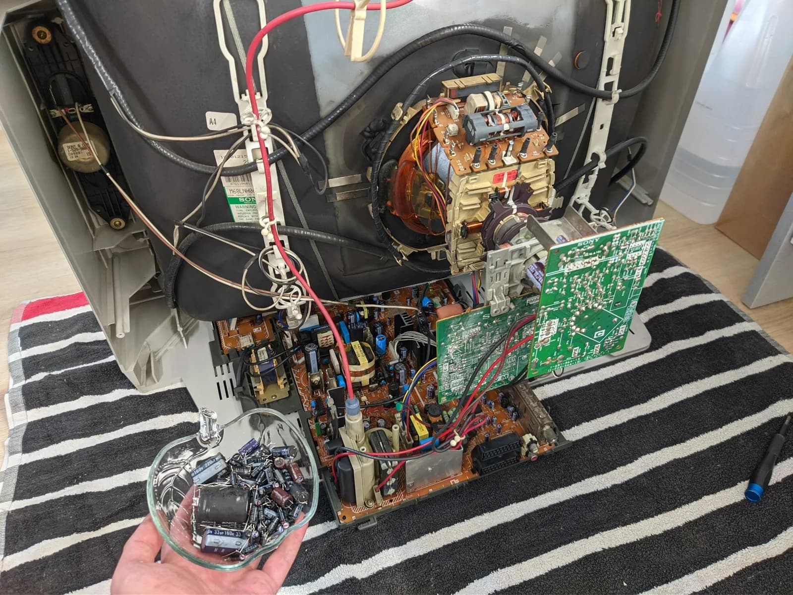 Deshelled Sony KV-29FX30E seen from the rear, the tube neck with its deflection yoke and red anode lead, the main chassis board still mounted (on this TV the PSU section and the main circuitry share a single PCB), and a hand holding a clear tub of the old capacitors pulled during the recap.