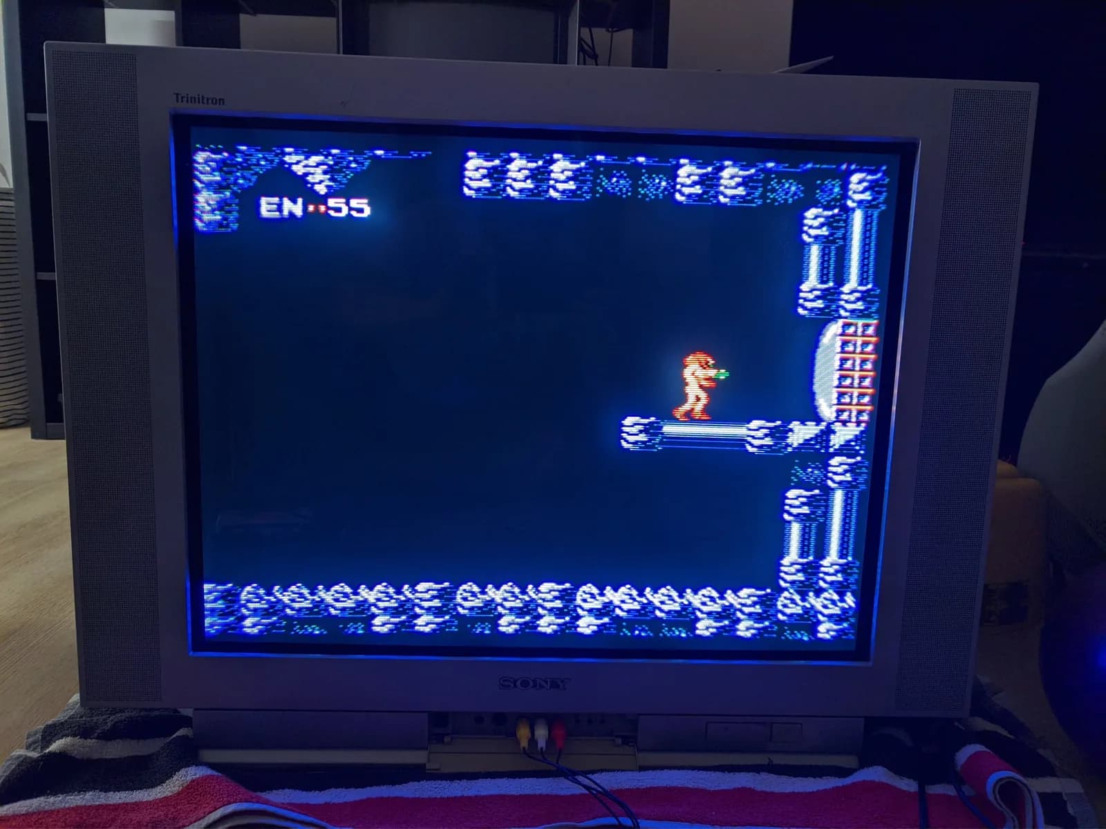 Reassembled silver Sony KV-29FX30E FD Trinitron TV running Metroid, Samus standing on a platform in a blue cavern with the energy HUD visible, composite cables plugged into the front jacks, sitting on a striped beach towel.