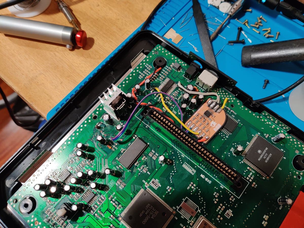 Soldered Mega Drive 2 RGB bypass mod installed on green PCB with colored wires, test setup on workbench with tools and components visible.