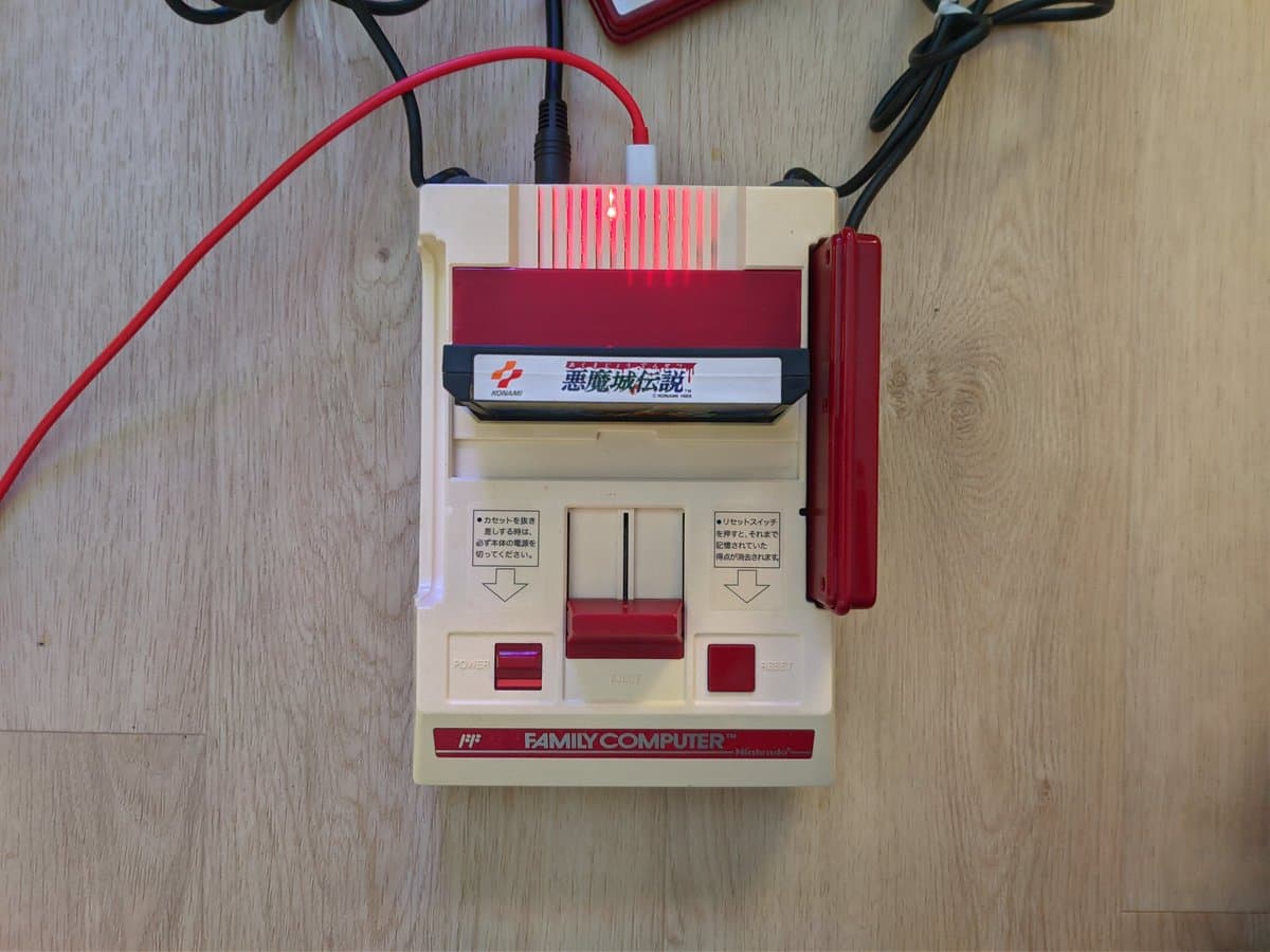 Nintendo Famicom with composite mod board installed, new red/cream stickers, power and reset buttons visible, connected cables on wooden surface.