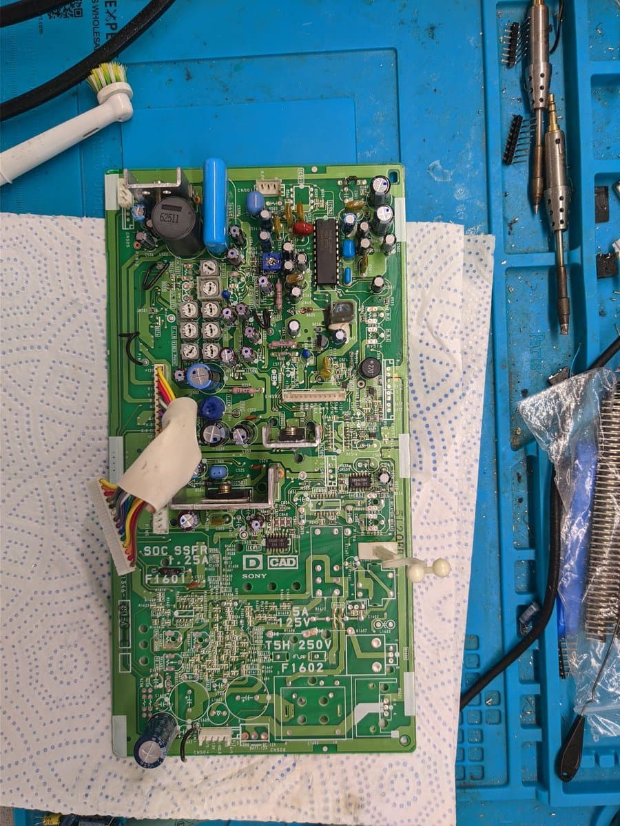 Sony PVM-9040 deflection board being cleaned with brush during repair, showing capacitors, transistors, and IC chips on blue work mat.