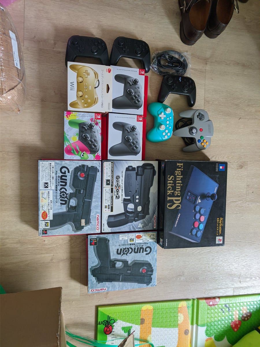 Flat lay of gaming controllers and light guns on wooden surface: black Pro Controllers, turquoise wireless controller, gray controller, boxed Wii light guns, fight stick, and game boxes arranged for documentation.