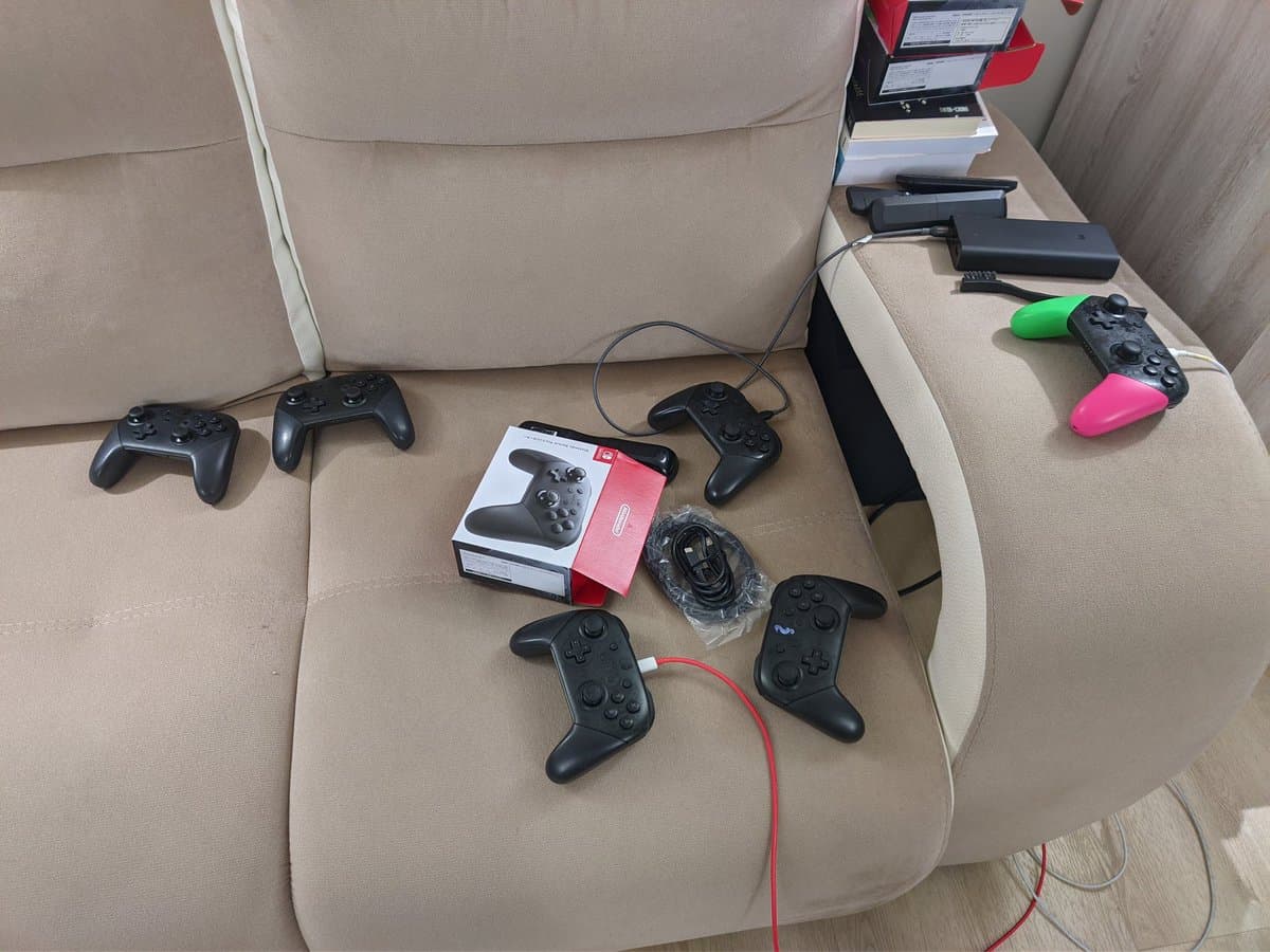 Six black Nintendo Switch Pro controllers arranged on a beige couch, with boxed controller and charging cables visible on the armrest behind them.