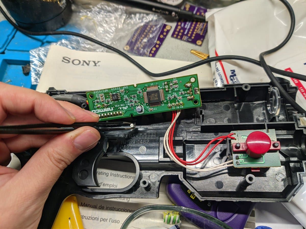 Hand holding green PCB with SMD components during GunCon light gun repair, black housing and red button visible below.