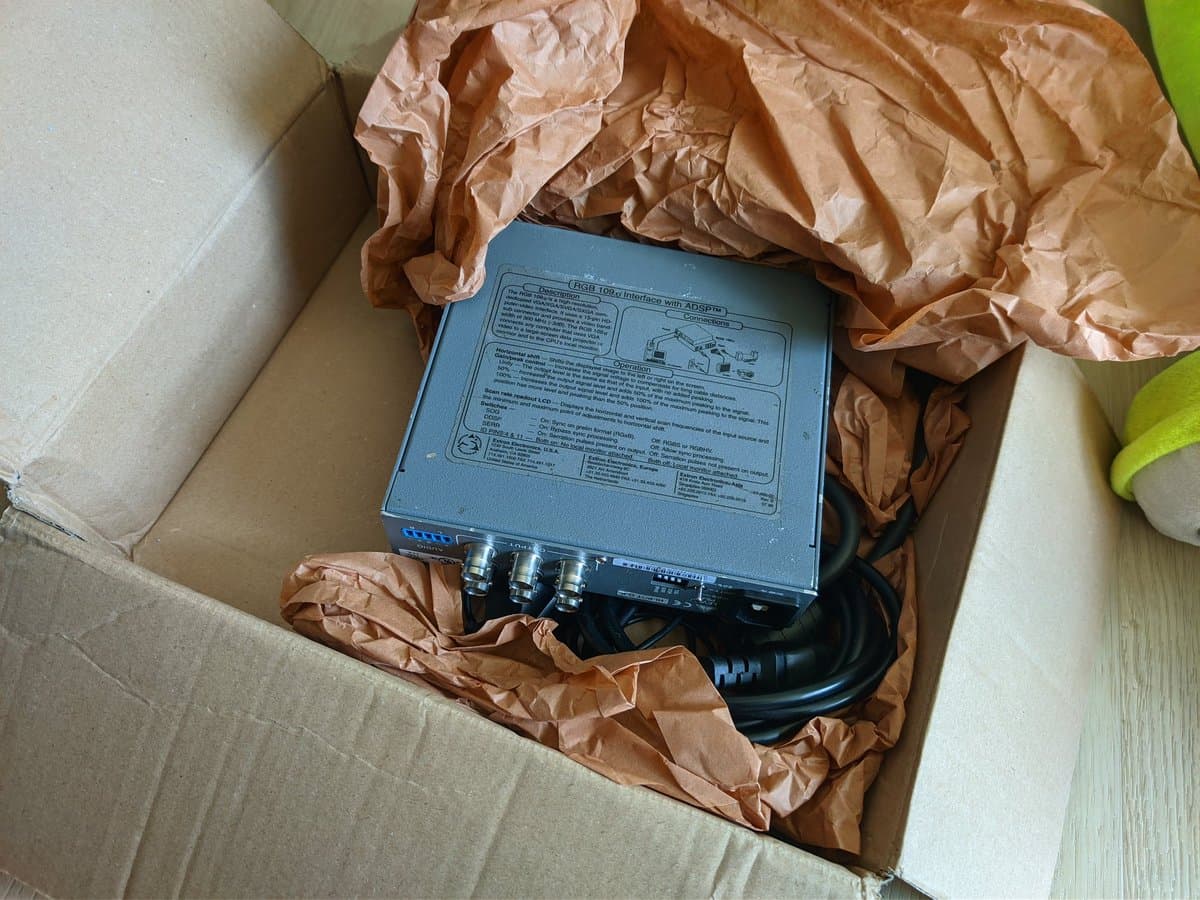Extron RGB 109xi scaler in brown kraft paper packaging inside cardboard box, showing blue device with three BNC connectors and power cable.