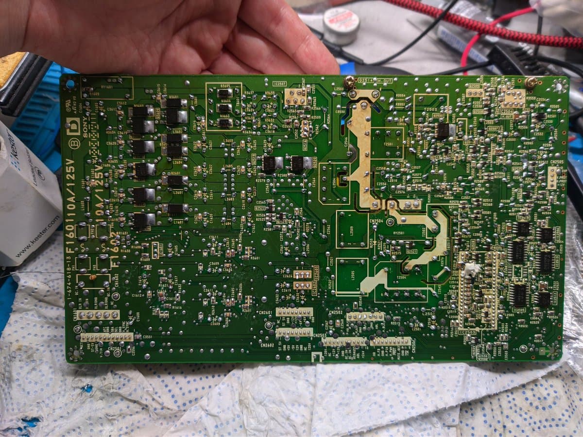 Sony BVM-D9H5J power supply board being held up, showing reflowed solder joints and messy flux residue from previous rework repair attempt.