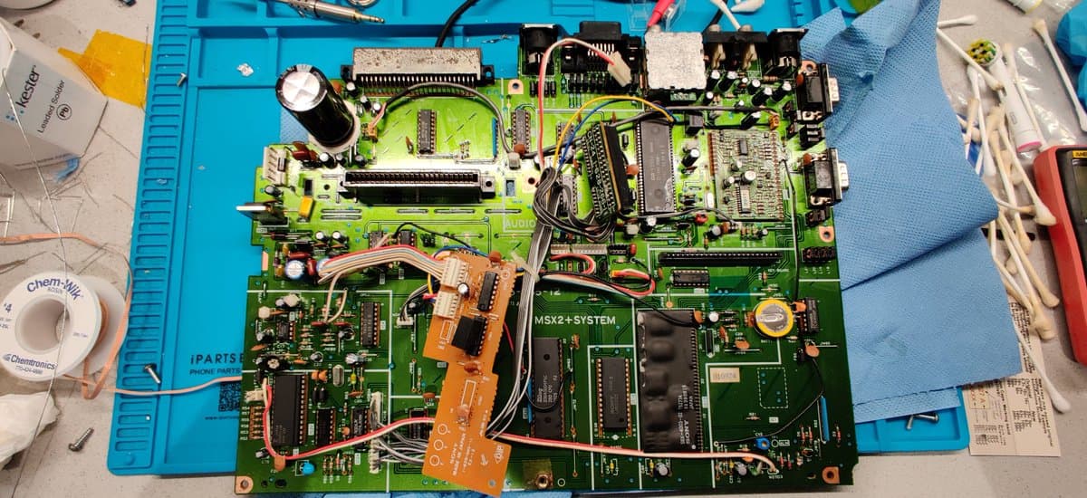 Sony HB-F1XV MSX2+ mainboard with replaced HIC-1 MLCC capacitors, orange daughterboard, and various ICs on blue work mat during restoration.