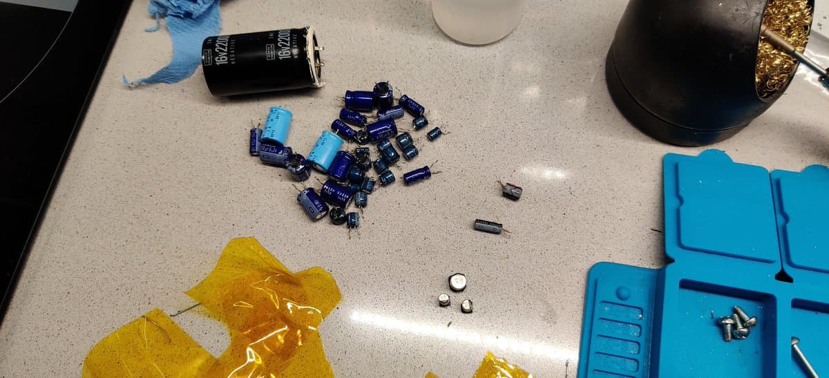 Disassembled HIC-1 board components including blue capacitors, small SMD parts, and a large electrolytic capacitor on workbench during MSX restoration.