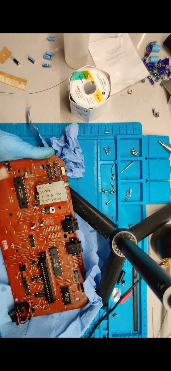 Sega SG-1000 II red PCB during recap with soldering iron, blue organizer holding capacitors and components, spool of solder visible above.