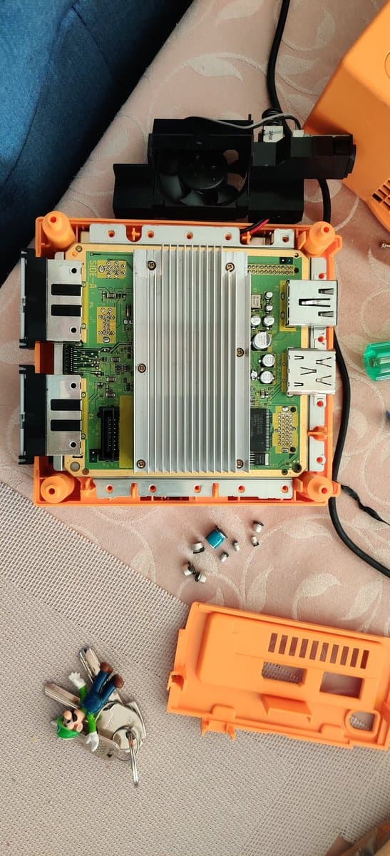 Disassembled GameCube motherboard with recapped components, orange housing, and Xeno modchip alongside desoldered parts and tools on work surface.