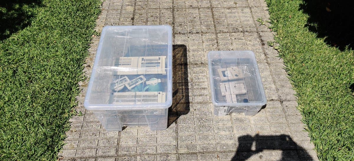 Two clear plastic storage containers on brick pavement holding Sega Mark III console parts during retrobrighting refurbishment process.