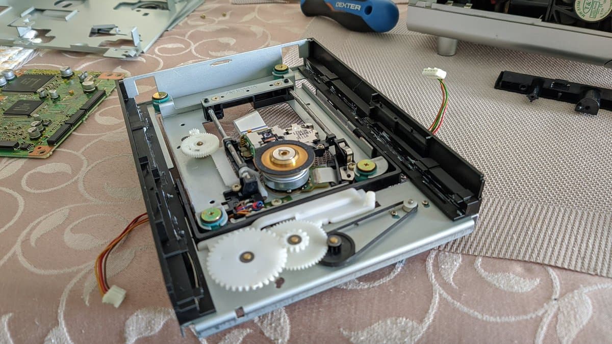Panasonic Q optical drive disassembled showing broken drive rail pieces, replacement 3D-printed rails, spindle motor with copper center, and internal gear mechanisms during restoration.