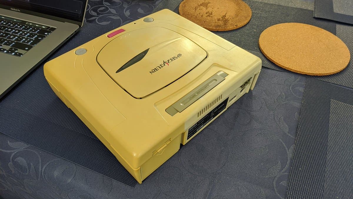 Yellow Sega Saturn model 2 with open disc lid, surrounded by cork coasters and laptop, on patterned desk mat during restoration work.
