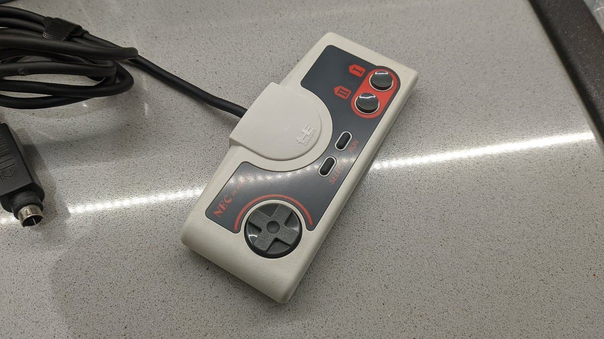 Retrobrighted Sega controller with red and black accents on metallic surface, alongside coiled cable and power connector.