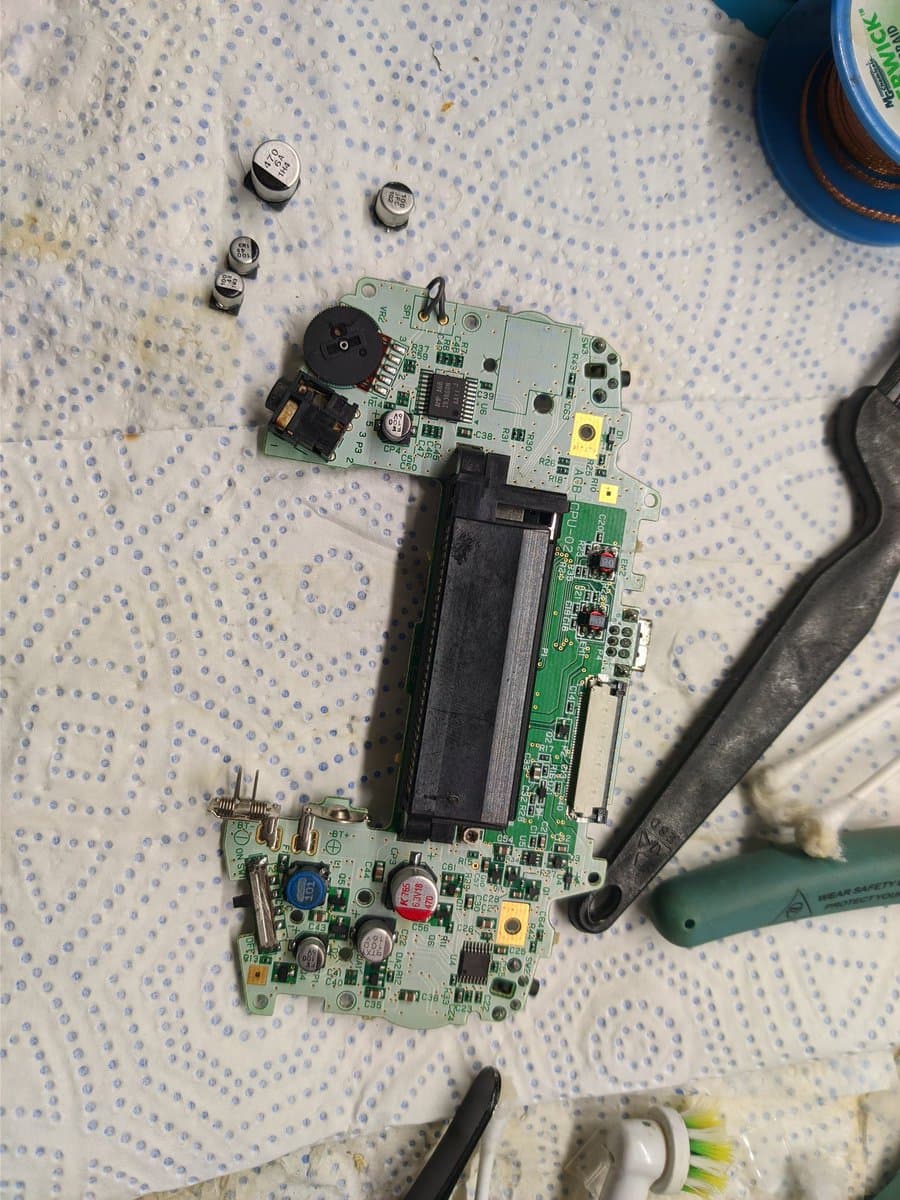 Disassembled GBA showing recapped motherboard with new electrolytic capacitors, removed battery cover, and desoldering tools on work surface.