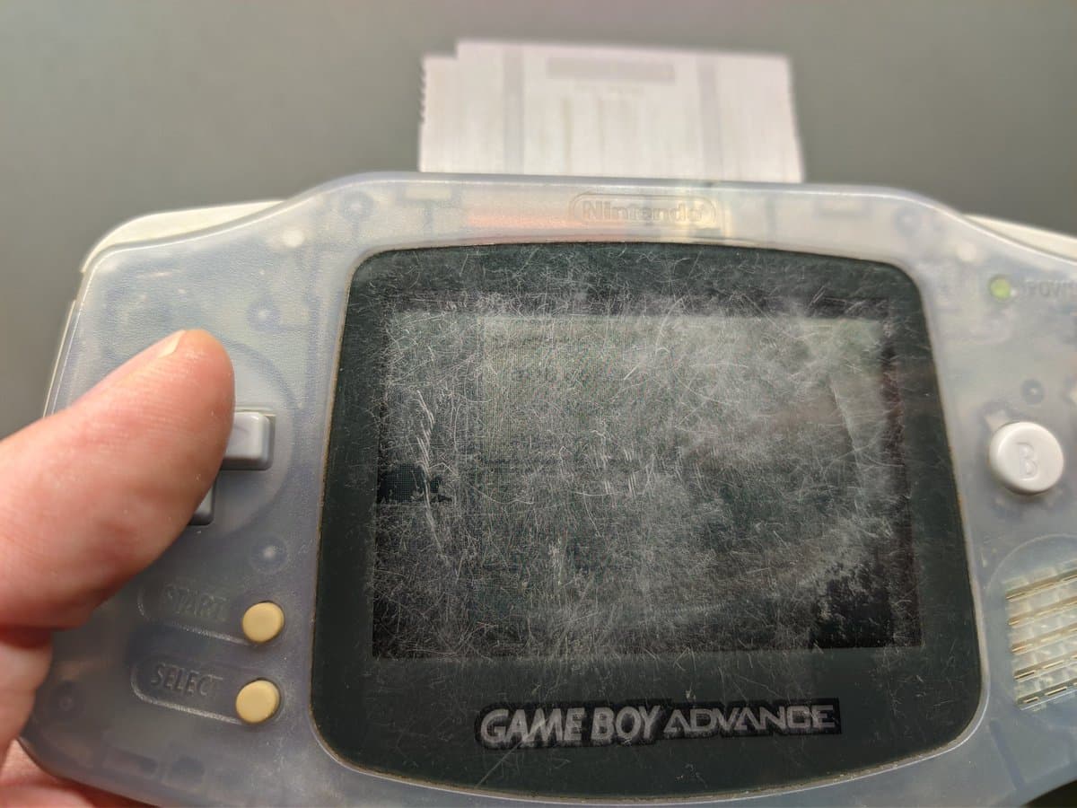 Heavily scratched Game Boy Advance screen held in hand, showing worn translucent shell and deteriorated display bezel before restoration work.