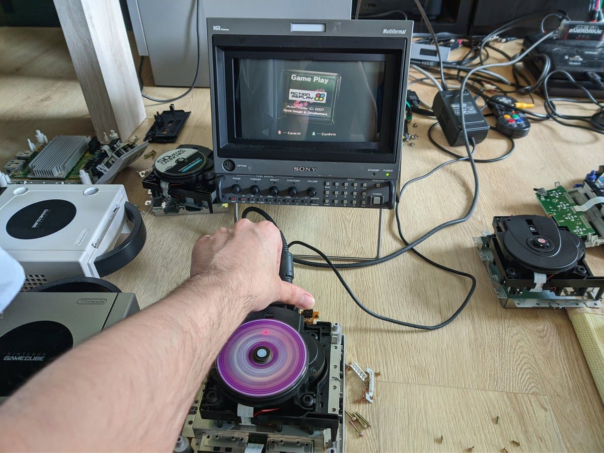 GameCube optical drive with spinning purple disc being held during capacitor replacement repair, Sony monitor displaying game in background.