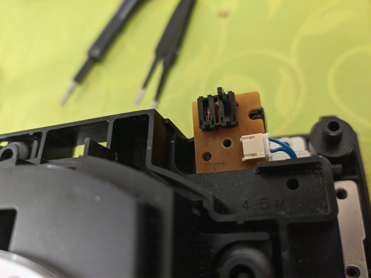 Close-up of GameCube optical drive PCB showing recapped components, white connector, and tweezers on yellow work surface with tools above.