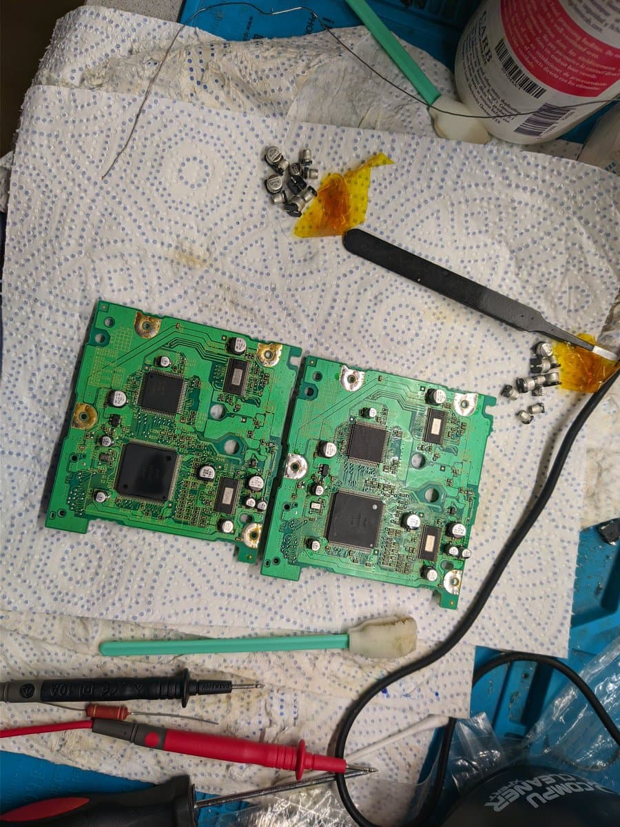 Two GameCube optical drive PCBs undergoing recapping repair, with desoldering tools, capacitors, and multimeter probes visible measuring motor resistance.