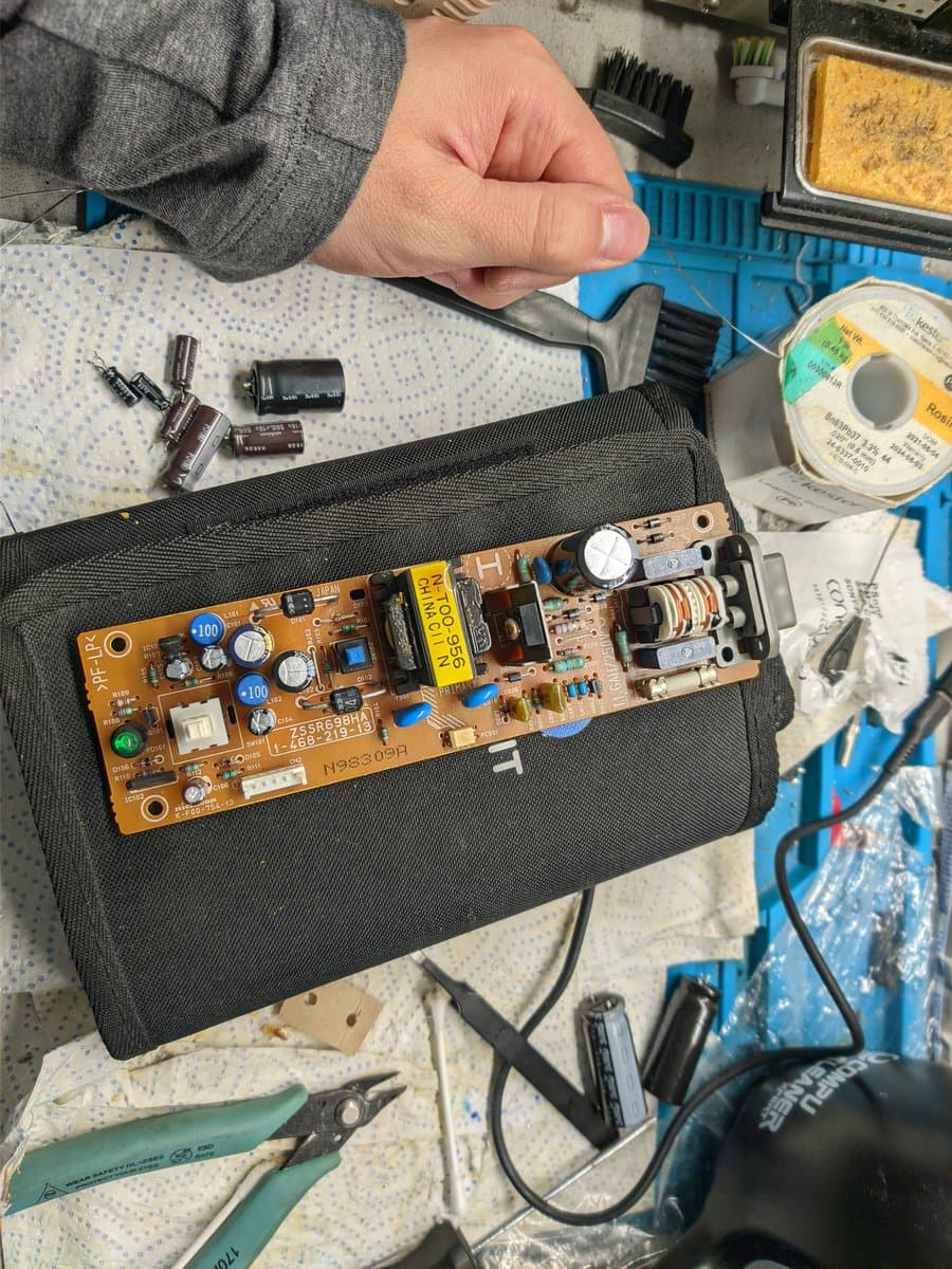 Technician holding open Sony PS1 PAL PSU board showing capacitors, transistors, and transformer with cracked solder joints on power jack during recap repair.