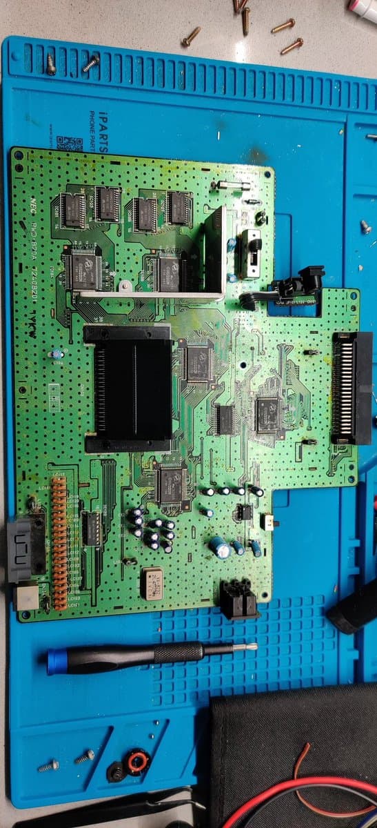 SuperGrafx mainboard on repair mat with new capacitors, voltage regulator, and soldering iron; scattered screws and components visible above.