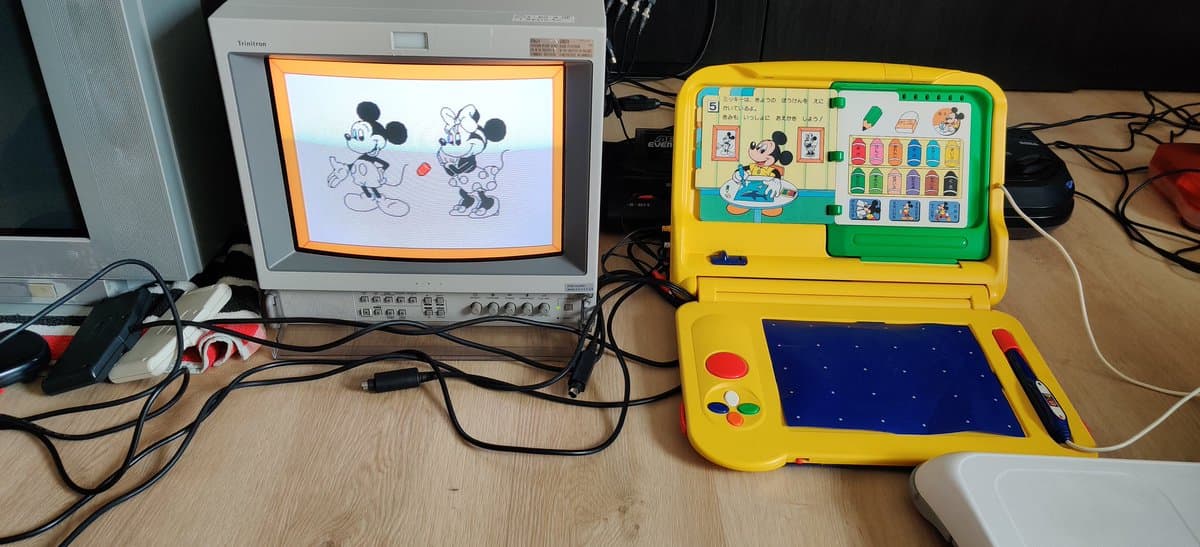 Recapped Sega Pico displaying Mickey Mouse game on CRT monitor; yellow handheld console with green cartridge slot showing game screen and controls.