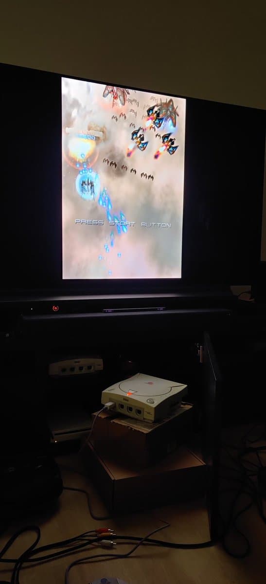 Sega Dreamcast VA0 displaying Ikaruga title screen via GDEMU mod, connected to display; console visible on shelf below.