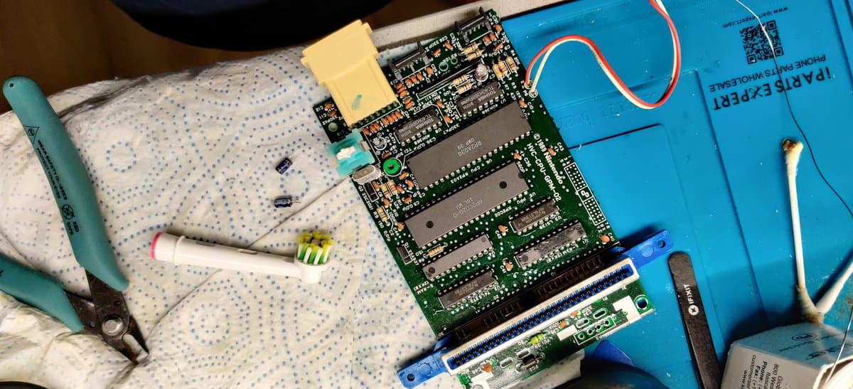 Famicom motherboard with new capacitors installed, composite mod board below, soldering iron, and testing components on work mat during repair and modding.