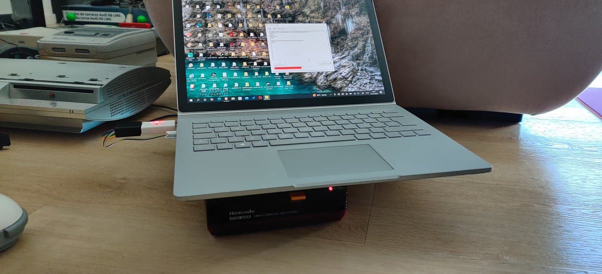 Homemade FDSStick cable positioned under silver laptop, black connector box with red indicator light visible, wooden desk workspace.