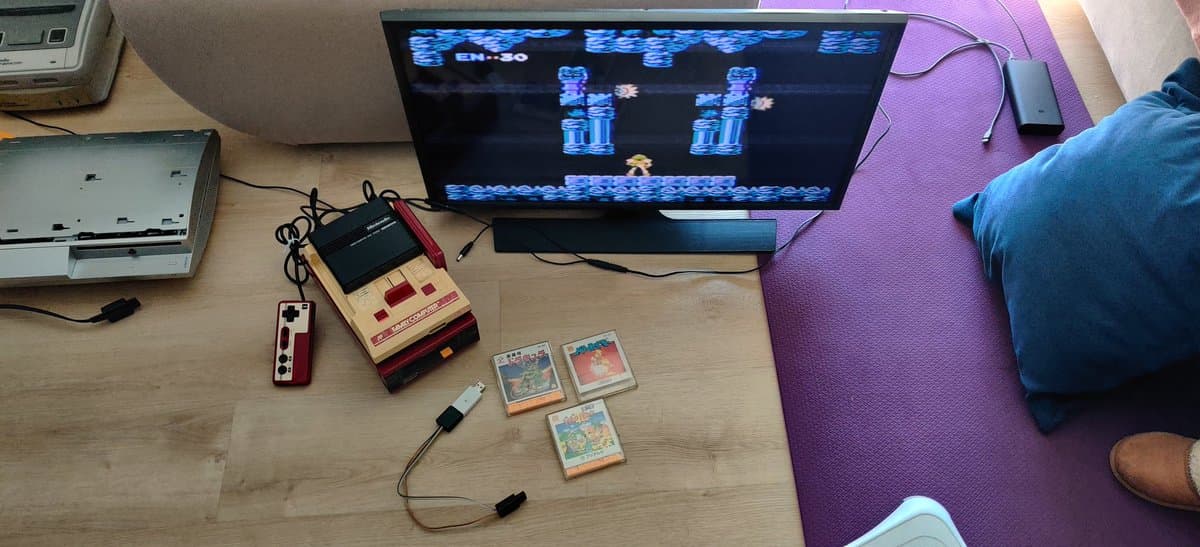 Famicom Disk System with homemade FDSStick transfer cable connected, three game cartridges displayed, retro gaming setup with monitor showing gameplay.