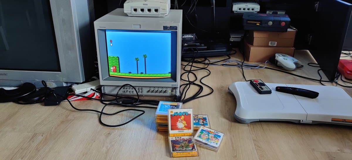 Famicom Disk System with CRT displaying game, FDSStick cable project, game boxes including Castlevania and Metroid on wooden desk.