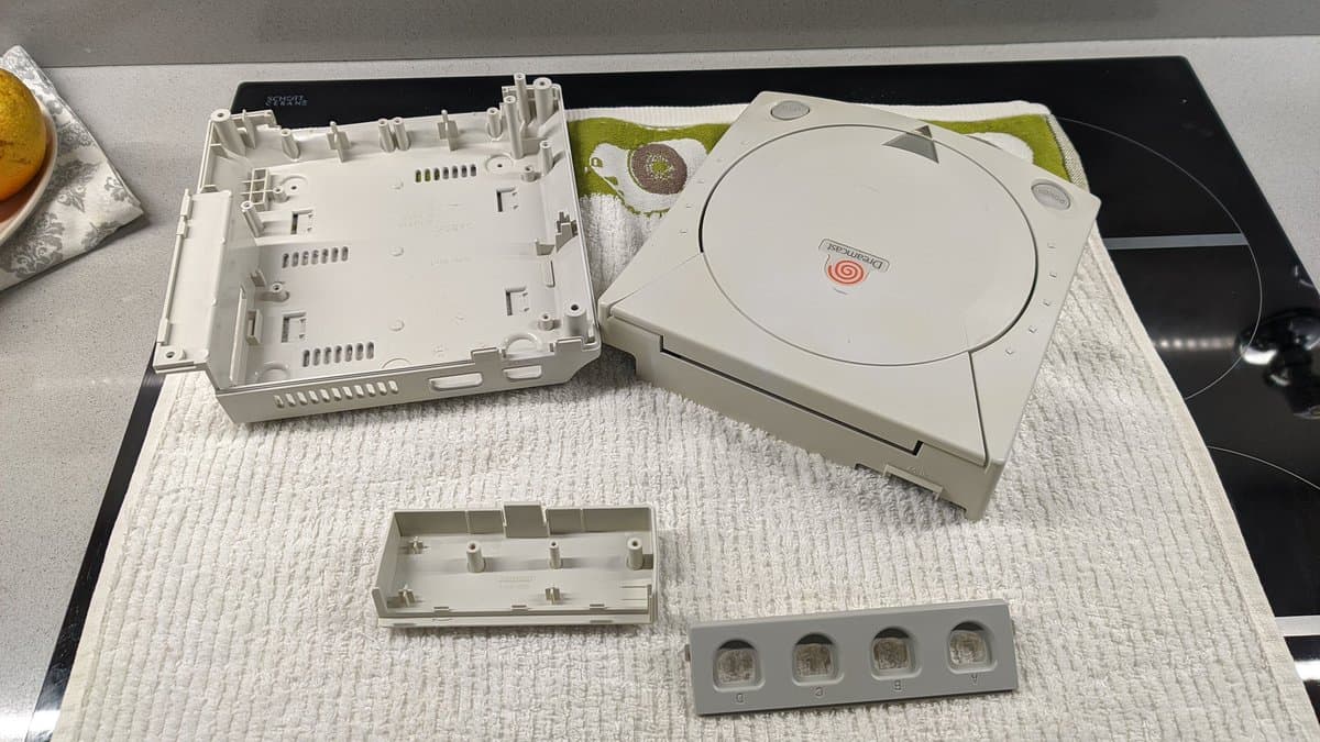 Disassembled Dreamcast VA0 components laid out for refurbishment: motherboard shell, disc drive mechanism, and front panel parts on work mat.