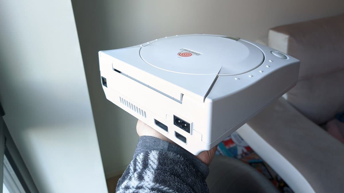White Sega Dreamcast VA0 console held in hand, showing disc drive, vents, power connector, and USB ports before refurbishment.