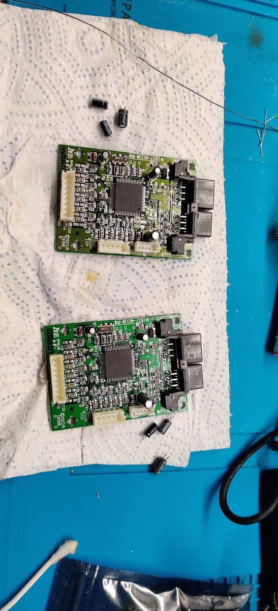 Two green circuit boards from Dreamcast arcade stick with removed capacitors on white anti-static mat during maintenance and repair work.