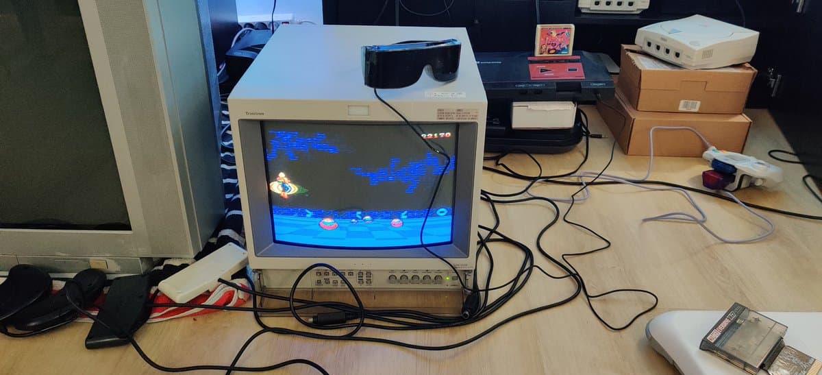Trinitron CRT displaying Space Harrier 3D with blue enemy sprites; Sega 3D glasses on monitor; Master System adapter and cartridge visible on desk.