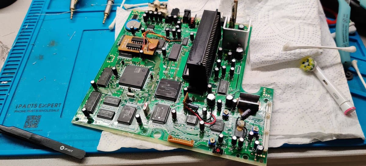 Sega Mega-CD 2 mainboard during recapping repair, showing replaced capacitors, IC chips, and heatsink with cleaning supplies on work mat.