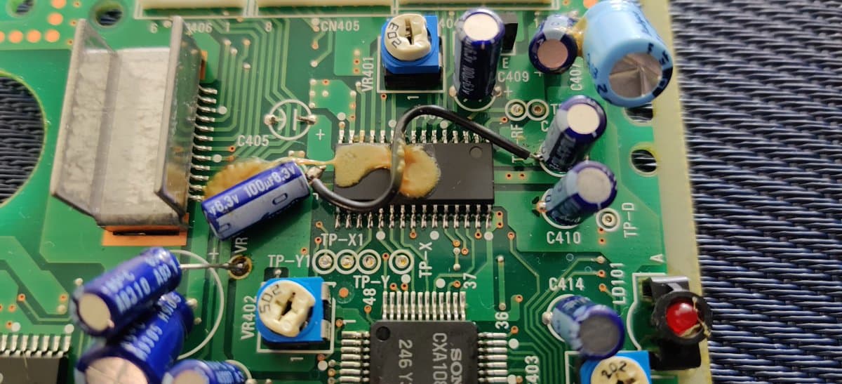 Sega Mega-CD 2 mainboard showing factory bodge wires, blown capacitors with tan-colored damage, and various ICs including SCN405 chip and blue capacitors marked 1000μ.