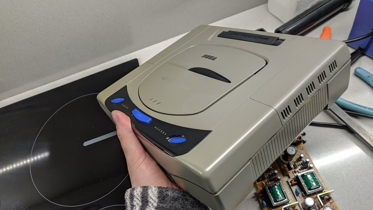 Hand holding beige Sega Saturn Model 1 shell during retrobrighting restoration, with circuit board and PSU components visible on workbench.