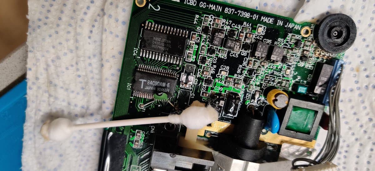 Sega Game Gear main PCB cleaned of electrolyte damage, showing exposed chips, capacitors, and solder joints with cotton swab applicator positioned on board.