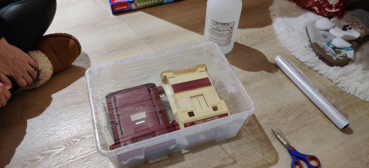 Famicom shells in clear storage container undergoing retrobrighting treatment, with chemical spray bottle, scissors, and restoration supplies on wooden work surface.