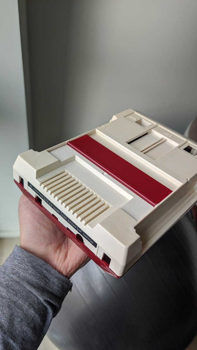 Repaired Famicom held in hand showing composite mod board installed, new red/white stickers, and internal components visible through open housing.