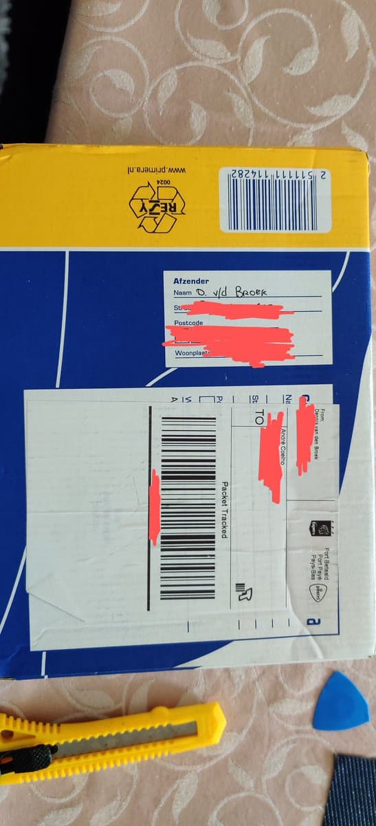 Disassembled PSOne motherboard with address labels redacted in red marker, barcode visible on yellow recycling bin above, craft tools nearby.