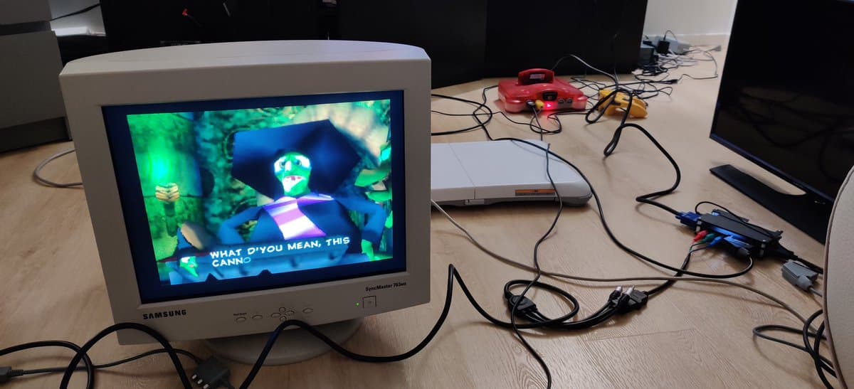 CRT monitor displaying retro console output, VGA cables and dongles visible on desk with various gaming hardware and testing equipment.