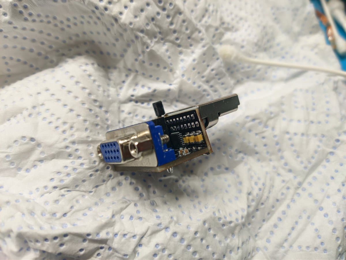 DC2VGA dongle on perforated mat showing blue PCB with IC chip, metal connector housing, and black plastic top cover.