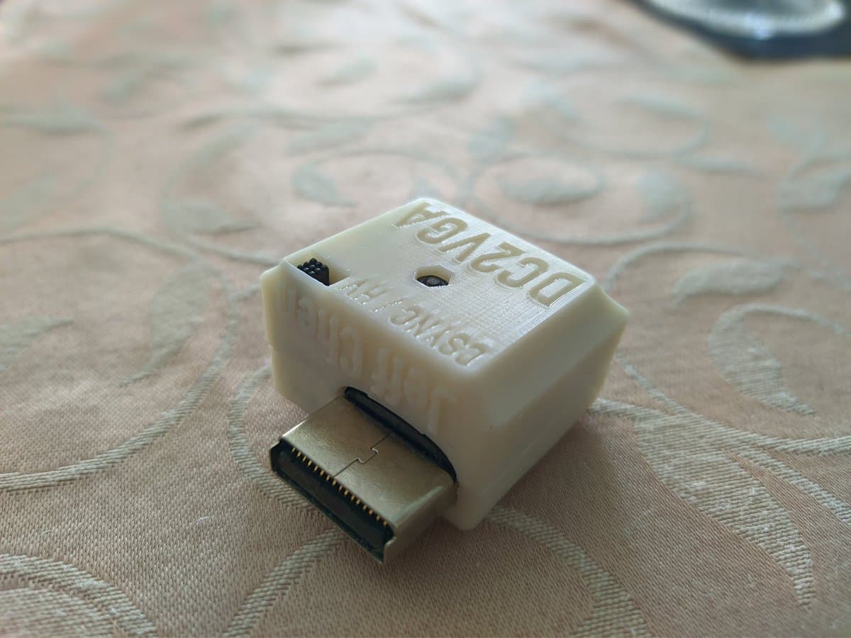 DC2VGA dongle by Jeff Chen for Dreamcast, cream-colored plastic housing with black connector port, displayed on patterned fabric surface.