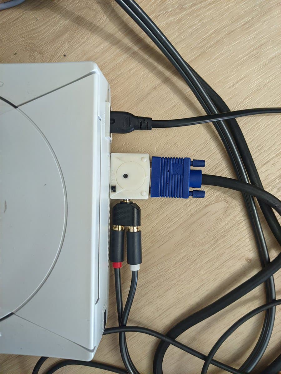 White console with VGA dongle connected, showing blue VGA connector, power cable, and audio RCA connectors on wooden surface.