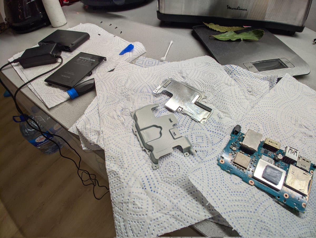 Disassembled Vita TV motherboard and shell on absorbent paper during cleaning and restoration, with various electronics and tools visible on workbench.