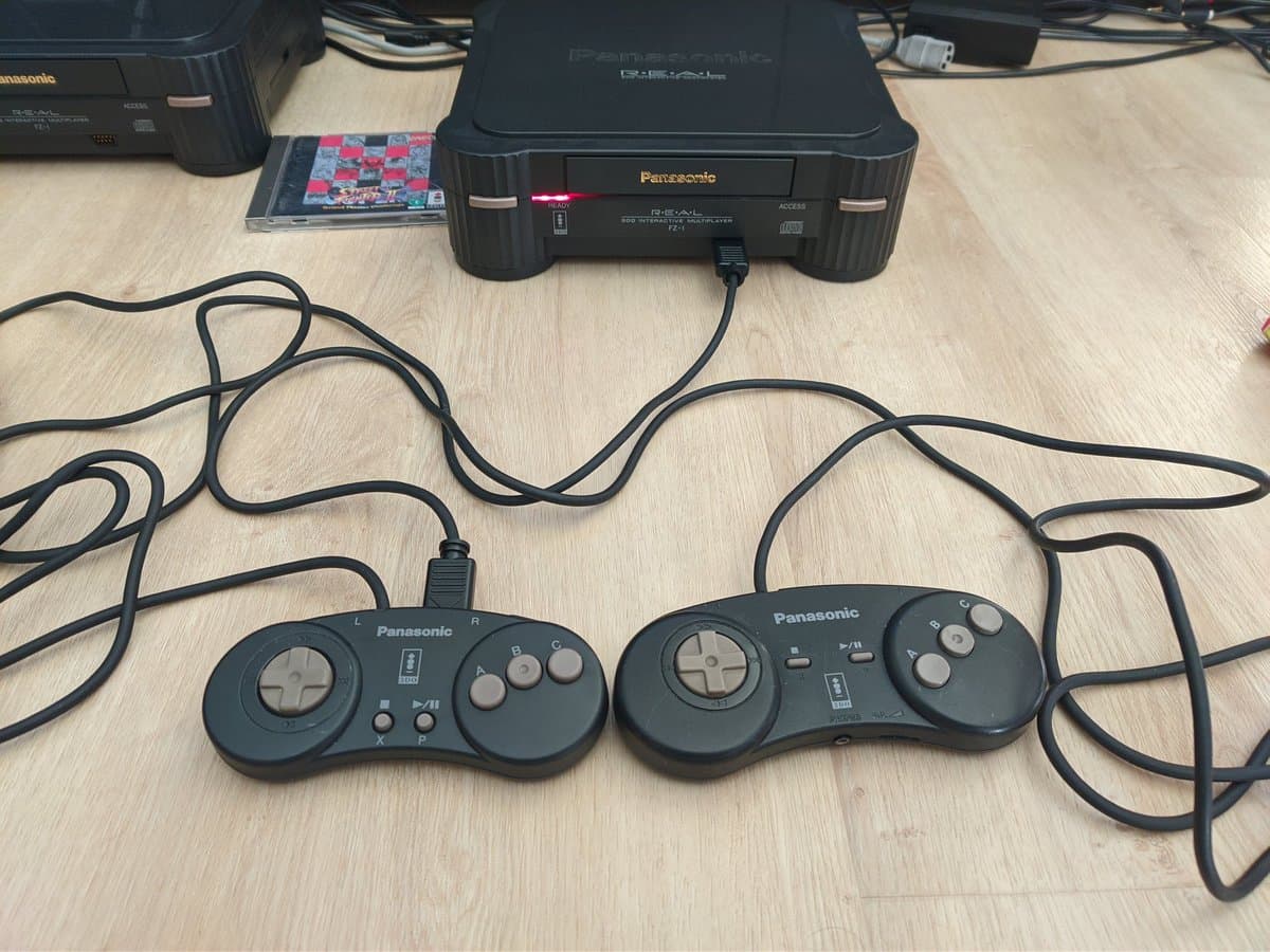 Panasonic 3DO FZ-1 console with two daisy-chained controllers connected via coiled cables, displaying S-video mod setup on wooden surface.