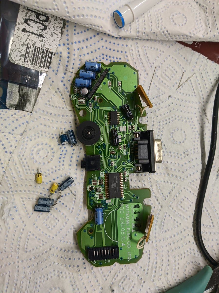 Disassembled 3DO controller PCB with removed capacitors laid out separately, blue tape, solder iron, and documentation during recapping repair.